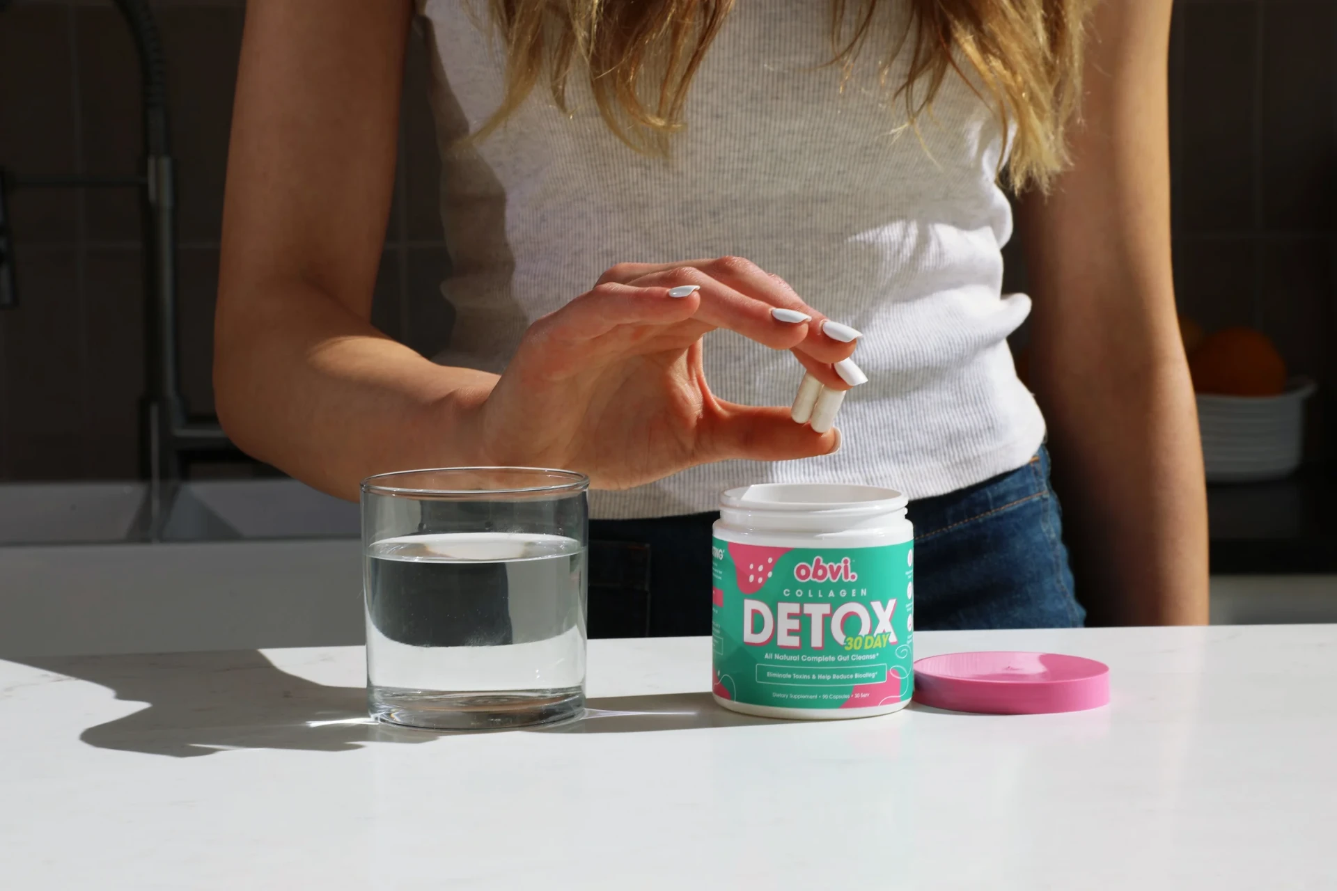 Three capsules held beside an open Obvi Collagen Detox jar and a full glass of water.